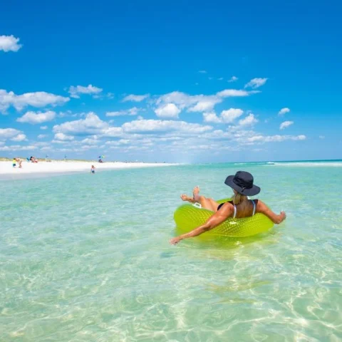 A woman relaxing on a lime green inner tube on the crystal clear water. There is a white sandy beach with people lounging in the distance. 