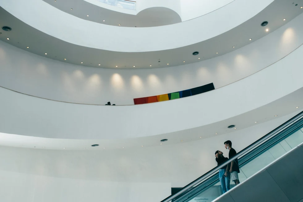 two people going up an escalator in a large room with white curved walls 