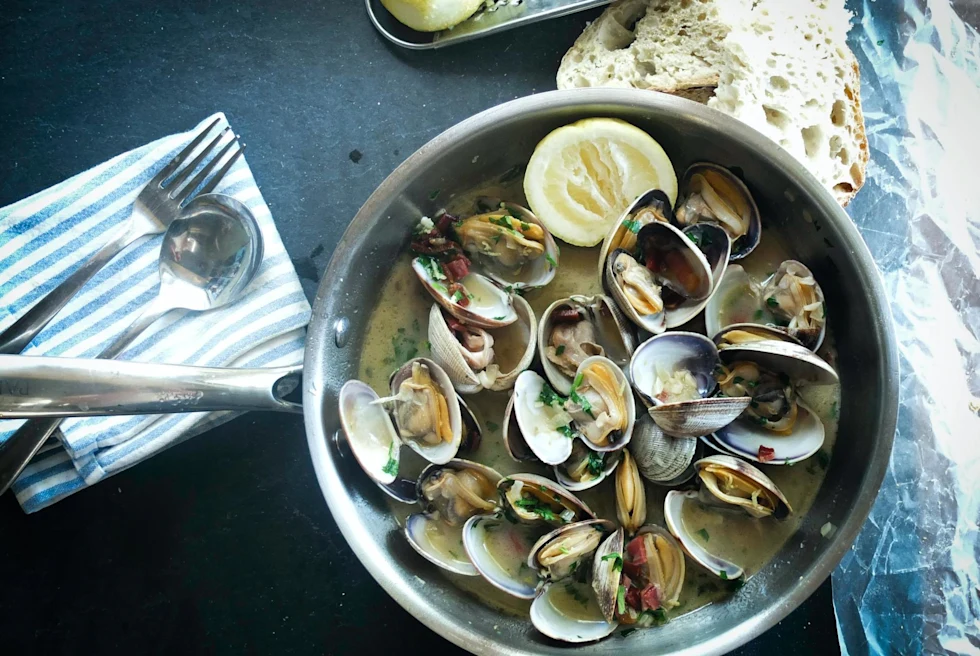 Mussels in broth with lemon with bread and utensils on the side on grey table.