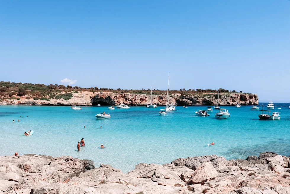 people wade in clear blue waters of an island's shallow beach with boats on a sunny day