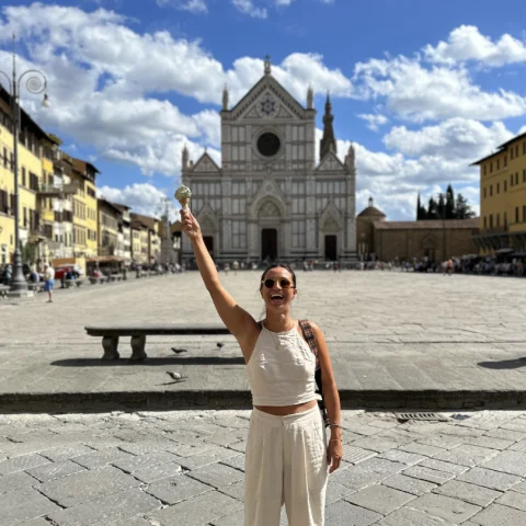An image of Janet wearing a white outfit and holding a gelato up in the air in front of a building in Florence, Italy.