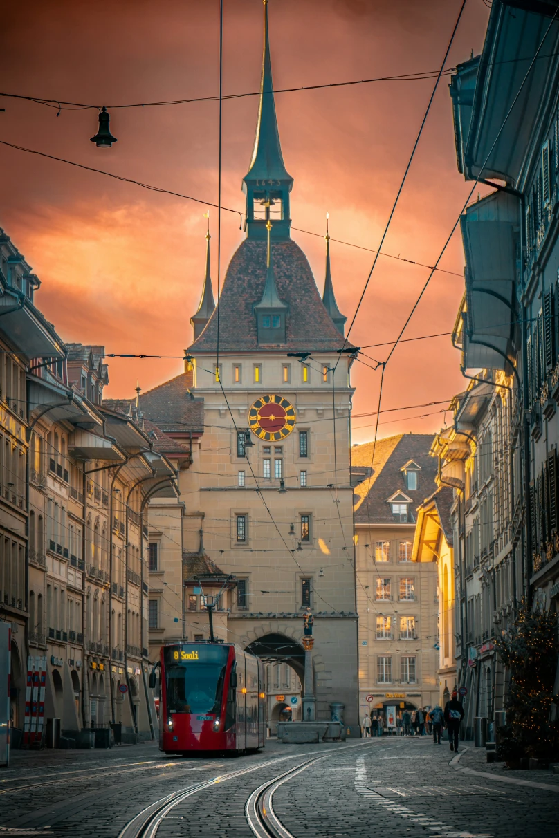 A red tram with a clock tower behind in Bern, Switzerland with an orange sky.