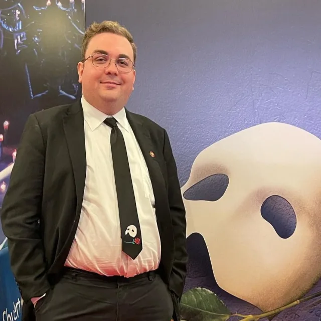 Travel Advisor Adam Landreneau in a black and white suite in front of a Phantom of the Opera poster.