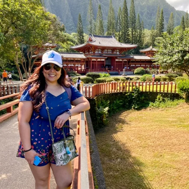 Travel Advisor Jodie Arcilla in a white hate and blue romper in front of a red temple with mountains.