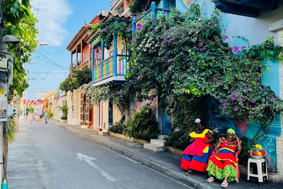 two women in colorful dresses sitting by a street lined with buildings and greenery