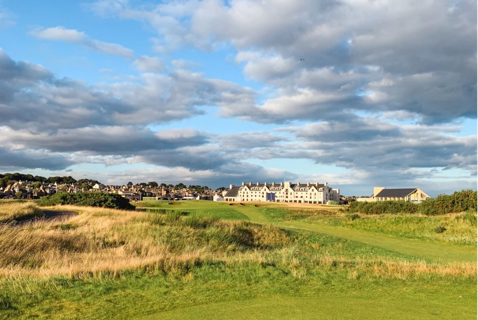 green and tan grassy golf course with white clubhouse building in distance and blue skies with white clouds