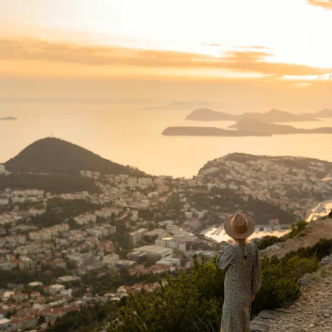 A girl in a hat and dress standing at a sunset on a hill in Croatia. 