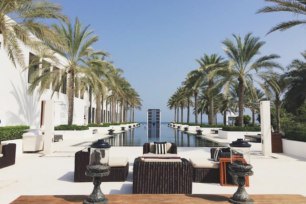 A view of a hotel pool deck with wicker furniture, a wood paneled deck, palm trees, black tables and a luxurious rectangular swimming pool down the middle. 