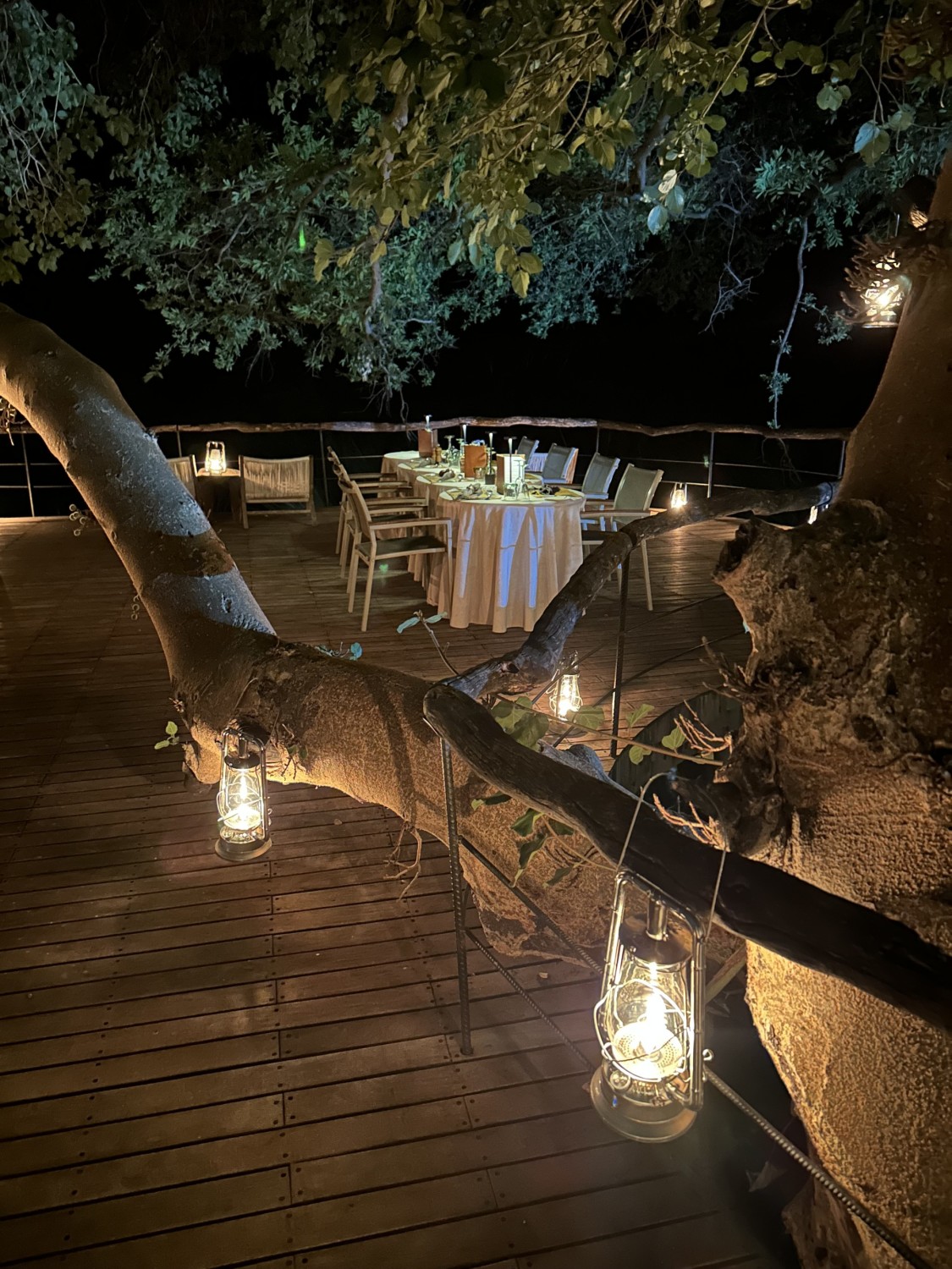 A table set up with a white tablecloth on an outdoor deck for a candlelight dinner under the stars with lanterns hanging from the trees at Sandibe