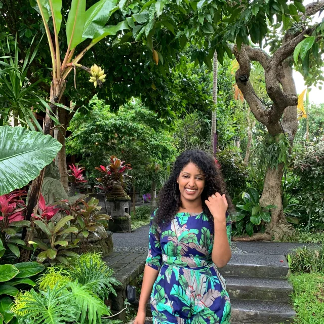 Picture of Bisaya in a floral print blue jumpsuit in a garden with tropical plants in the background