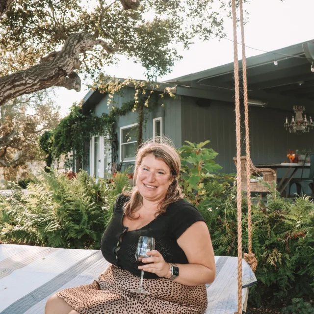Travel advisor Rachel Kantrowitz sits on a large swing holding a wine glass.
