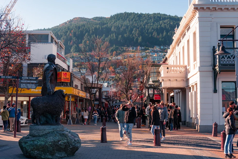 A busy town with a statue, people walking, buildings and trees covering a mountain in the distance.