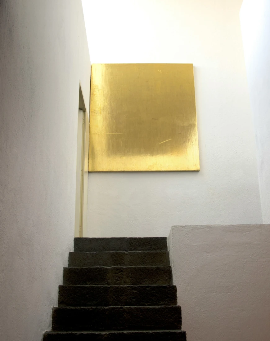 a staircase leads up to a gold square hanging on a sparse white wall