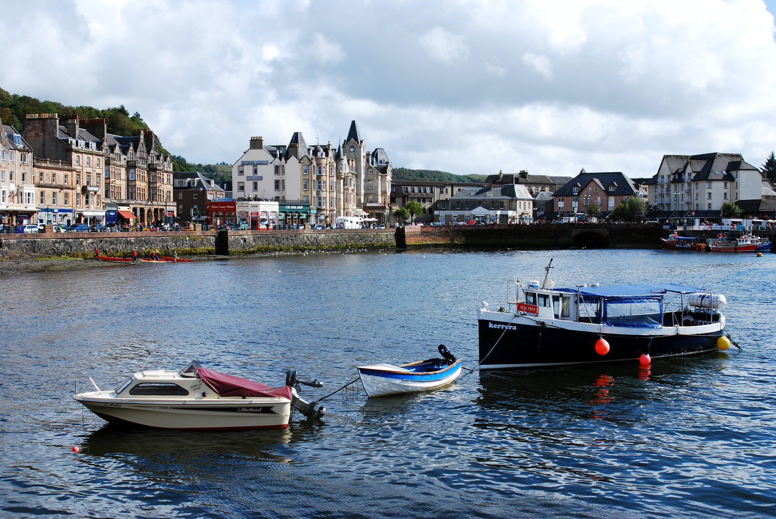 A blue lake with three white and blue boats floating and white and brown tall buildings around in Oban, Scotland.