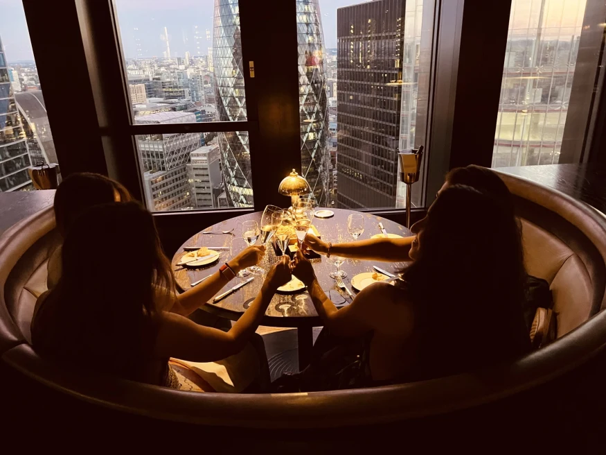 a group of 3 women clink their wine glasses at a round table with a view of the city through the windows 
