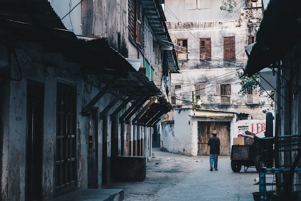 A dark shadowed alleyway with white buildings and a person with blue jeans black shirt and tan hat walking