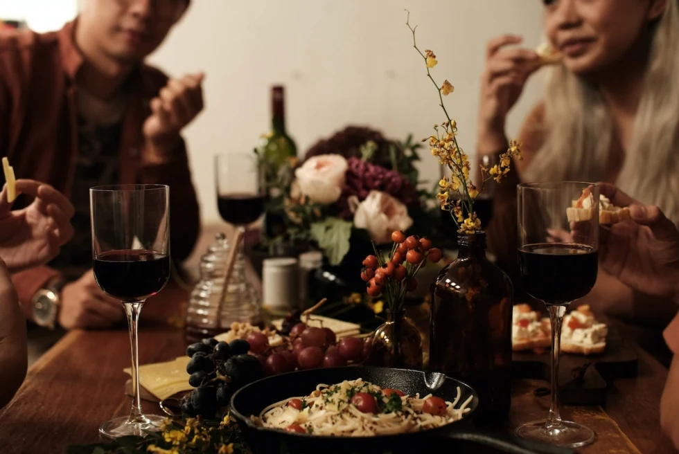 people sitting around a dinner table with flowers, pasta, and glasses filled with red wine