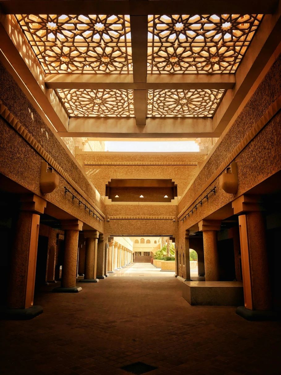 A picture taken inside of a corridor of a brown and white concrete building with a patterned roof. 