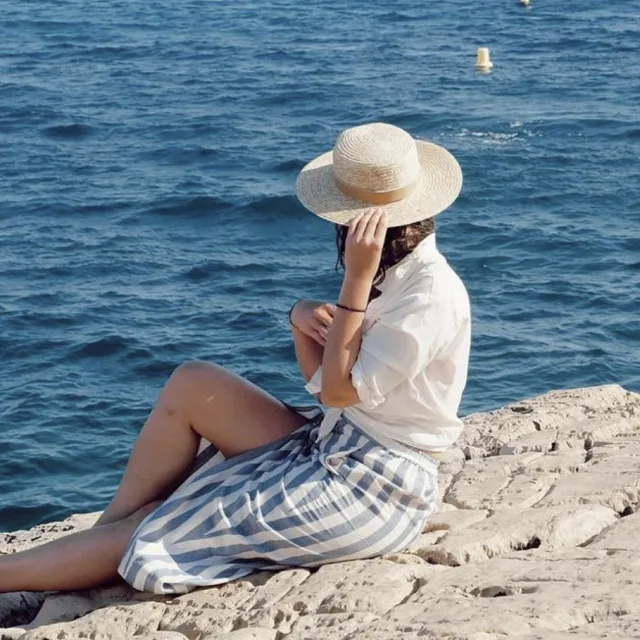 A picture of Amy wearing a white shirt and blue and white striped bottoms while sitting on stones with a sea view