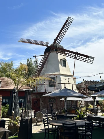 A windmill behind an outdoor patio with umbrellas, tables, chairs and string lights, as well as various plants. 