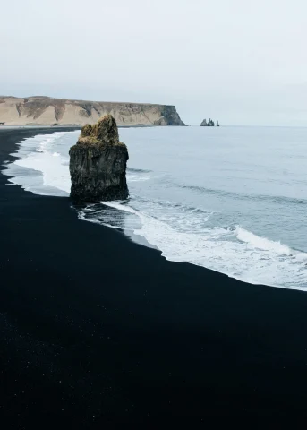 A picture of a black sand beach with waves rolling in on the shoreline. There is a large boulder in the middle of the scenery, and a rocky cliff in the background. 