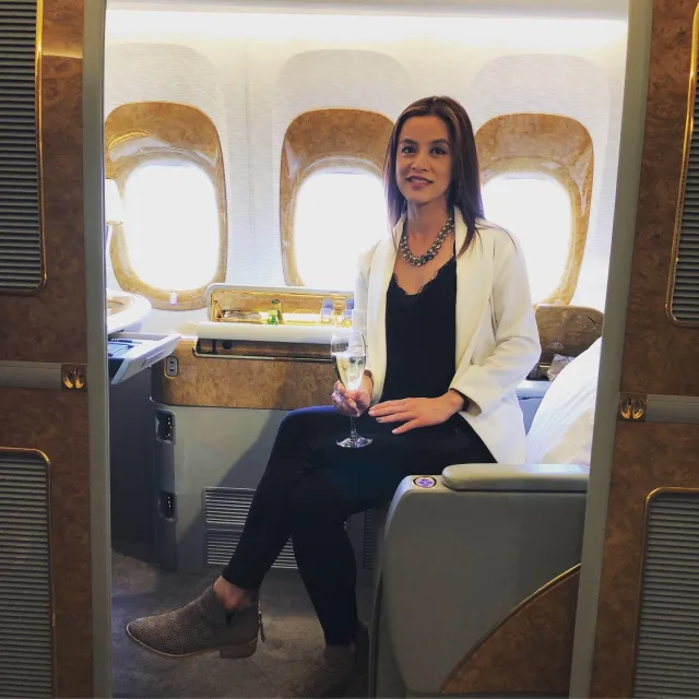 Travel Advisor Christina Trami Dang in a black shirt and white jacket sitting on an airplane with champagne.