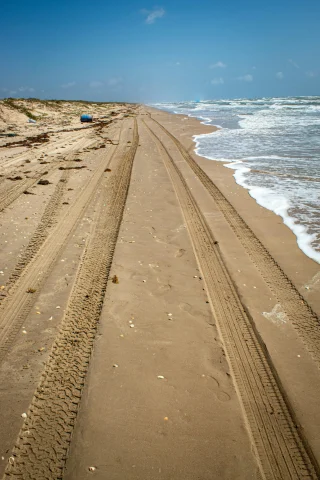 A picture of vehicle tires on seashore in the daytime on the beach. The ocean waves are rolling in on the right. 