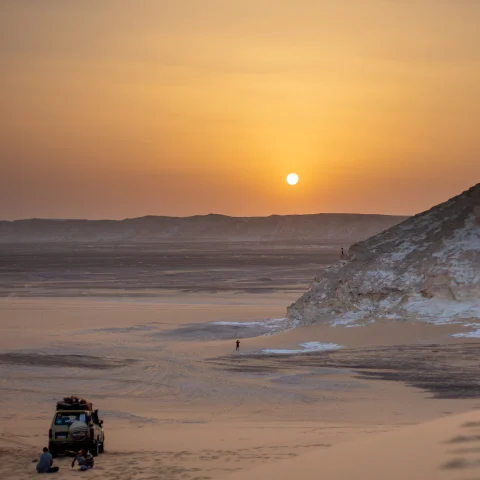 sunset over a calm desert with a car and a person walking towards the sun