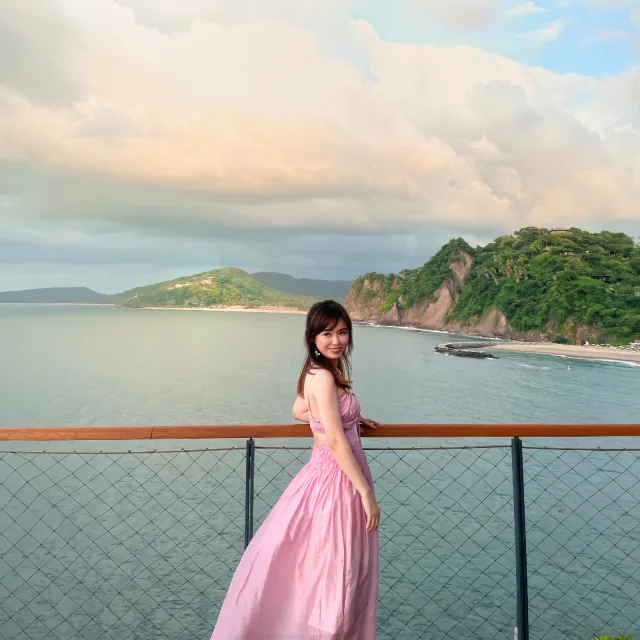 Jessica Zhou in a pink dress standing against a fence with the sea and mountains in the background