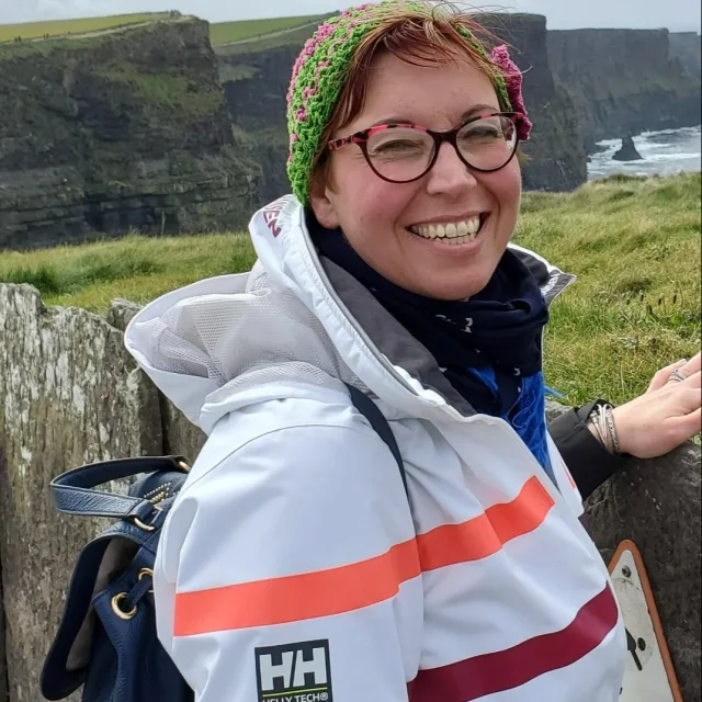 A picture of Susan wearing a striped white jacket and green and pink hat looking over the cliffs of Ireland