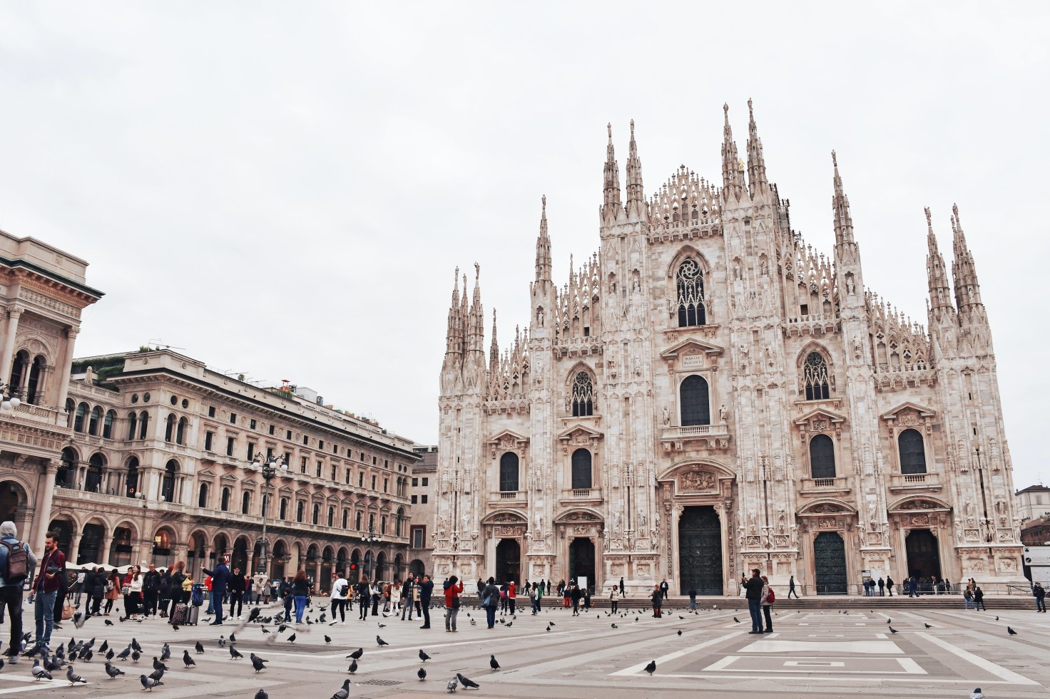 A white gothic church opening up to square with grey pigeons and tourists in black clothing at Duomo Cathedral Square in Milan, Italy.
