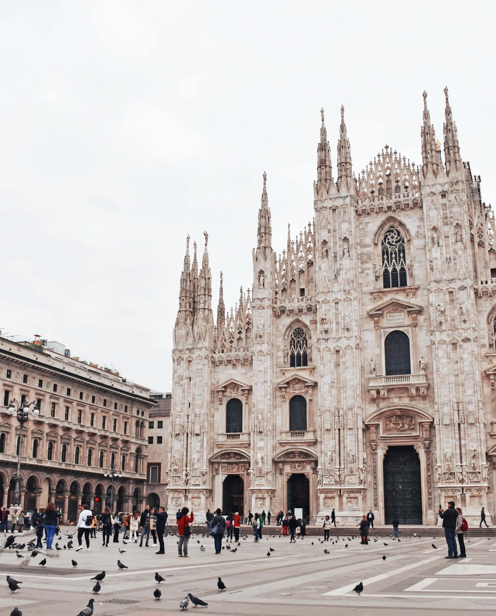 A white gothic church opening up to square with grey pigeons and tourists in black clothing at Duomo Cathedral Square in Milan, Italy.
