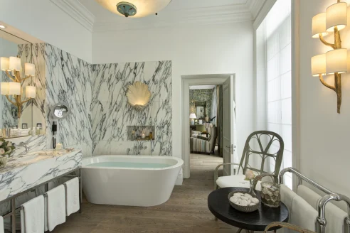 luxe bathroom with wooden floors, a large tub filled with water, wall lamps and marble wall tiles
