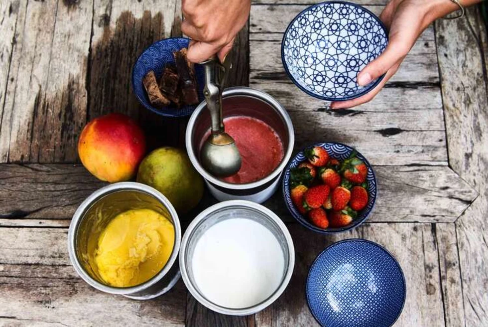 a hand scoops pink sorbet from a bowl with other sorbets and fresh fruits