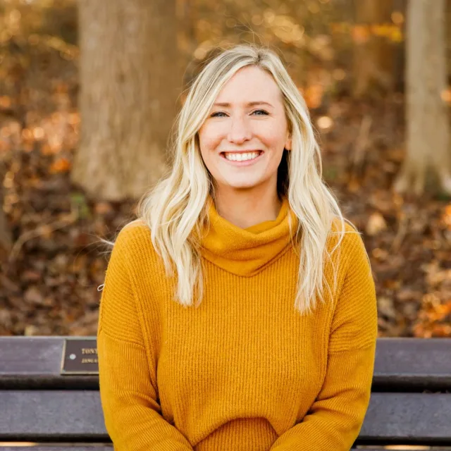 Travel advisor Kylie Dowling wears an orange sweater and sits on a park bench in the fall