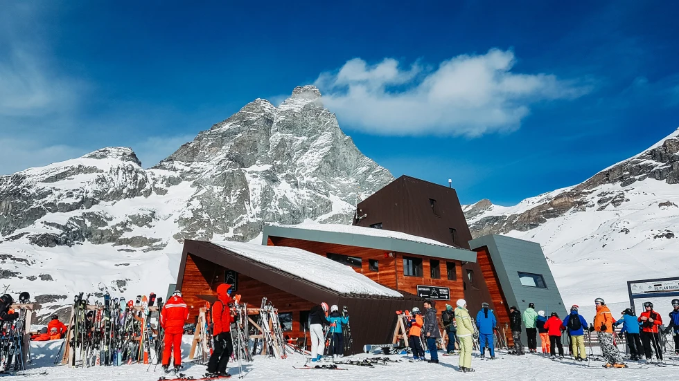 skiers standing outside building with large mountain in the background