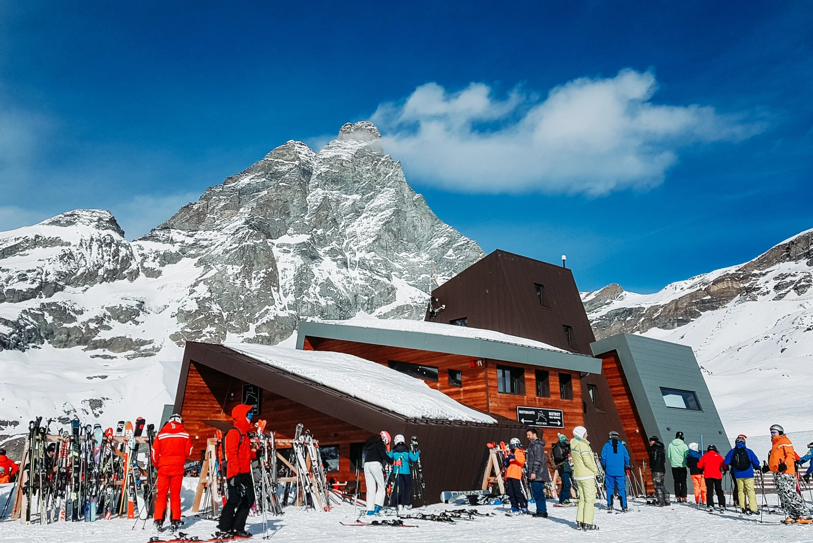 skiers standing outside building with large mountain in the background