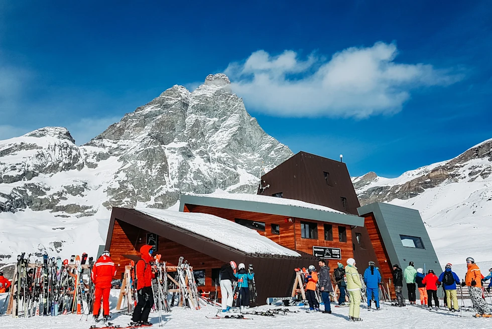 skiers standing outside building with large mountain in the background