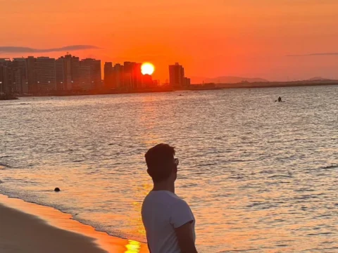 A man looking out to a beaming orange sunset peaking through the city skyline and shining onto the water from the beach. 