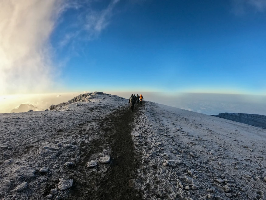 A few hikers in the distance trekking on a mountain with snow on the stone and a path down the middle overlooking a blue sky and overlook of white clouds