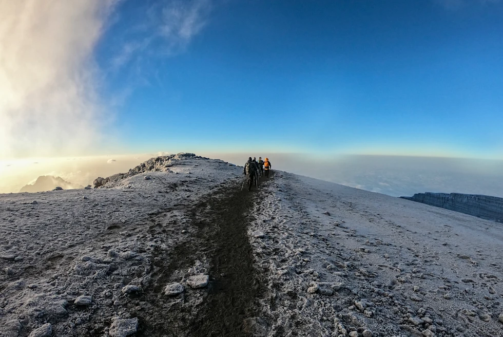 A few hikers in the distance trekking on a mountain with snow on the stone and a path down the middle overlooking a blue sky and overlook of white clouds