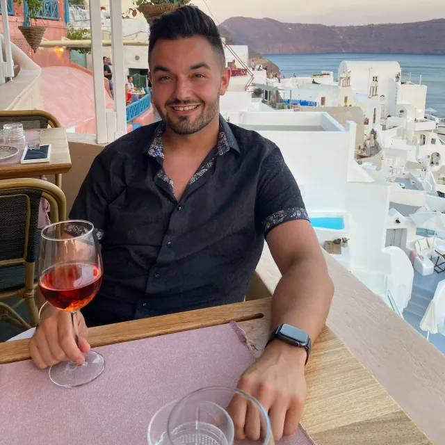 John wearing a black button up shirt and holding a glass of wine while seated at a wooden table with a view of white buildings, the sea and mountains in Greece in the background  