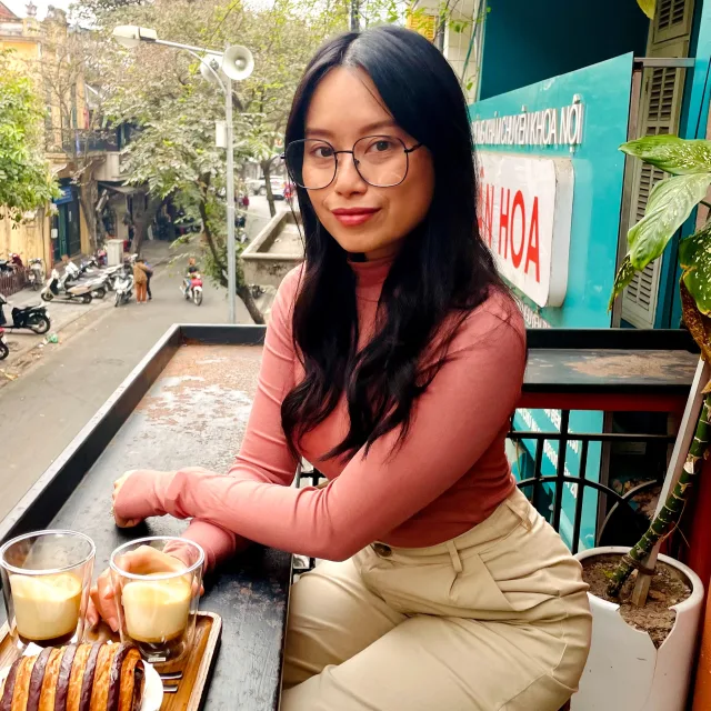 Cam Le in a pink top and khaki pants sitting at a balcony with two drinks and a pastry