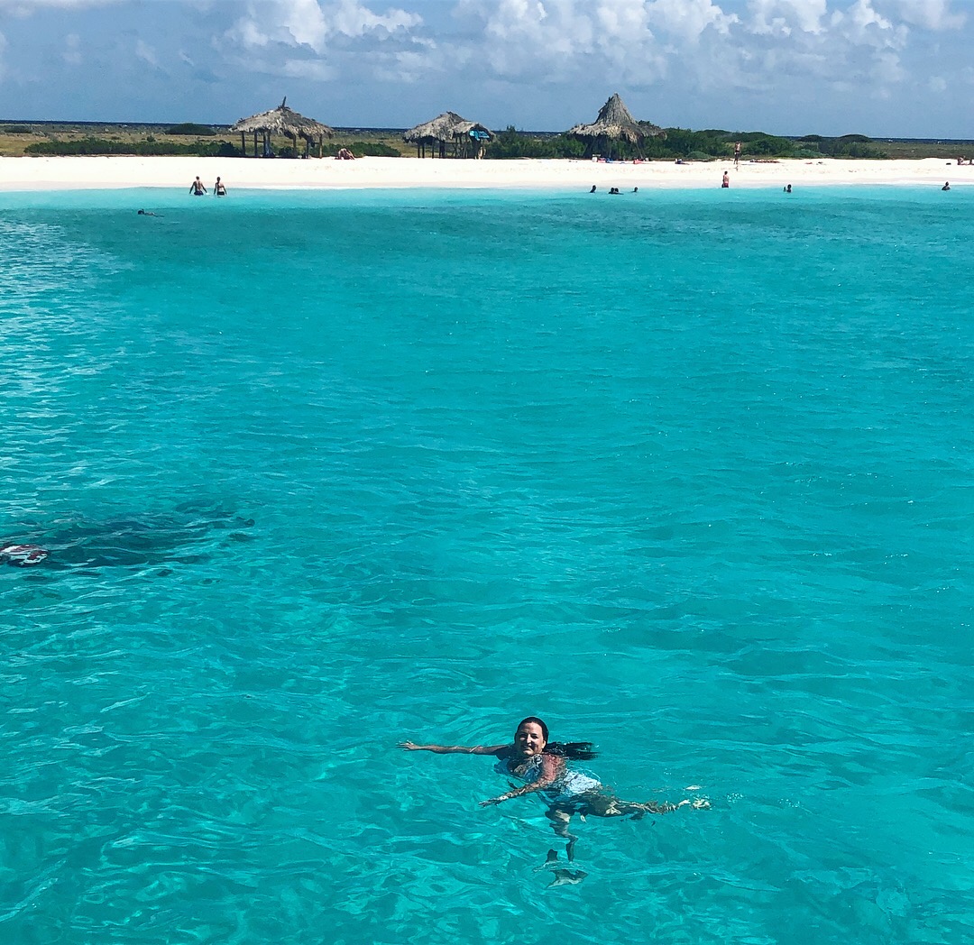 Samantha smiling and swimming in the azure waters of a tropical beach, with white sand and tiki style huts on the shore.