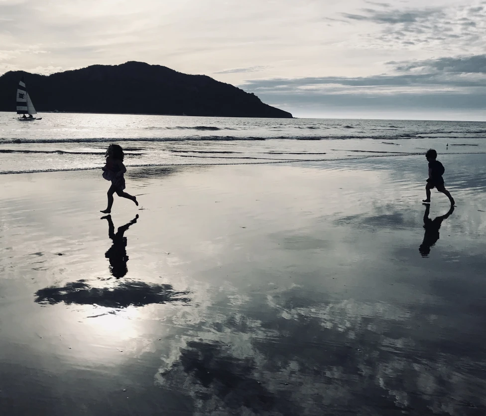 Two kids running on the beach with an ocean and a mountain in the background.