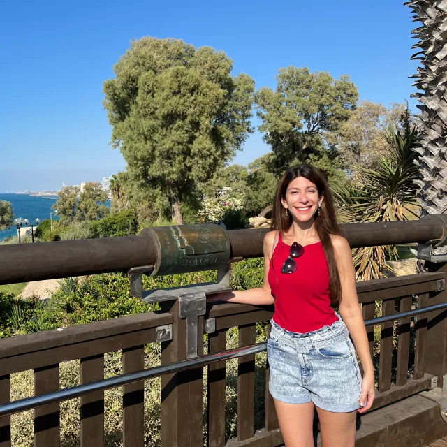 Travel Advisor Miri Paz in a red tank top and jean shorts in front of trees and the ocean.