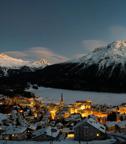 An elegant alpine resort town at nighttime surrounded by snowy mountains, water and lit up buildings.