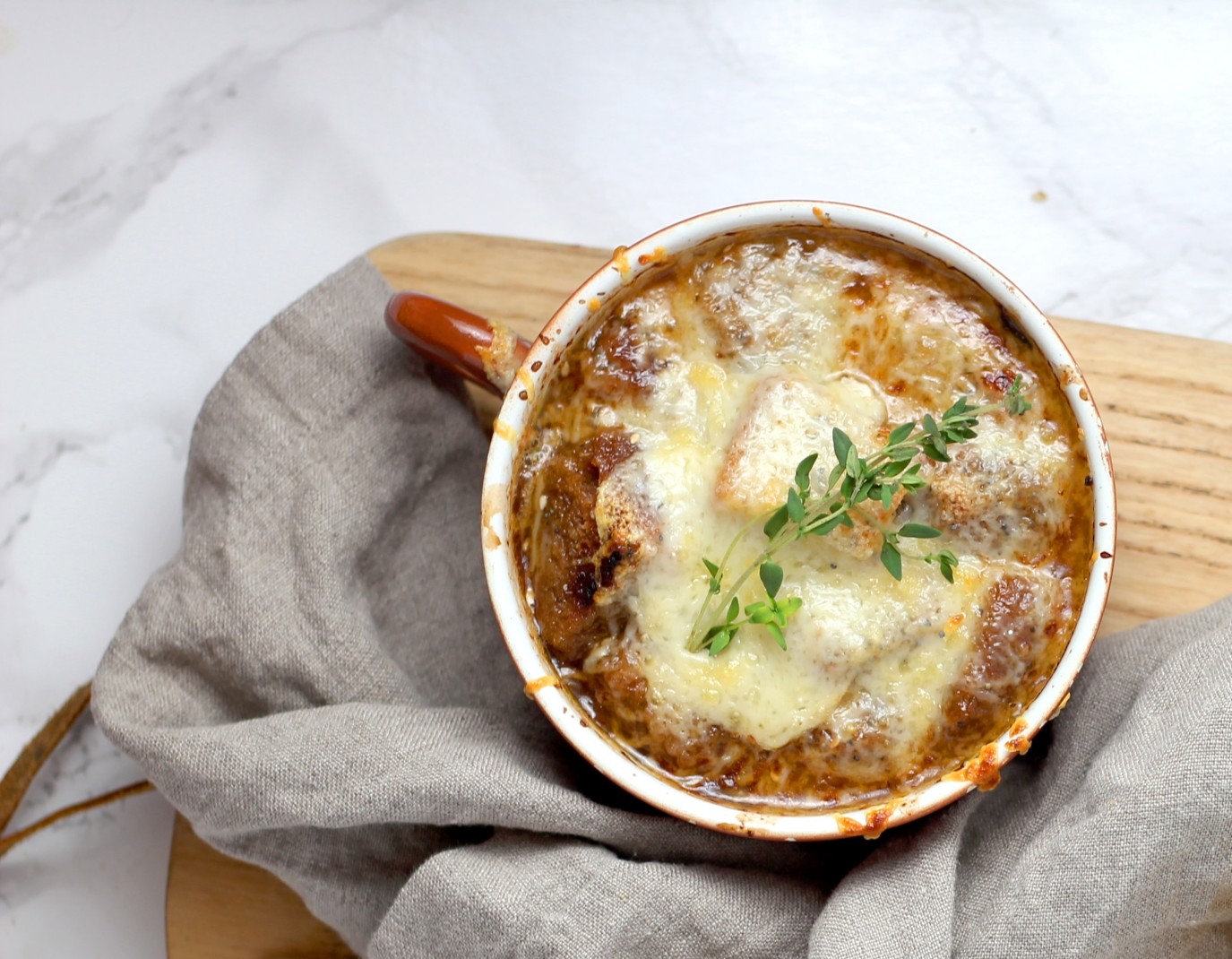 Hot onion soup in a mug topped with fresh herbs on a wooden cutting board