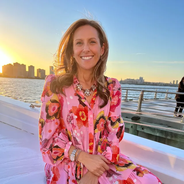 Tracy wearing a pink dress and sitting on a boat with the city skyline, sunset and water in the background