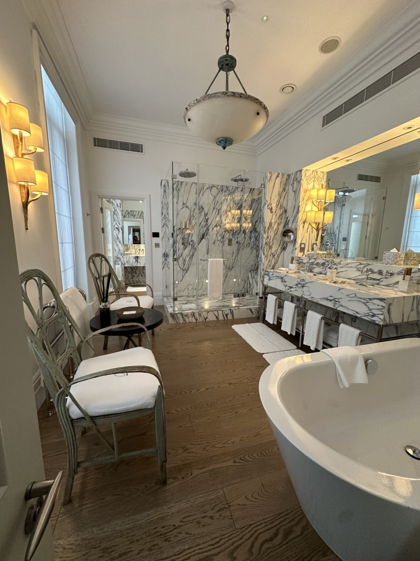 The bathroom in the Kipling Suite at Brown's hotel showing white marble tiles, a large hanging ceiling lamp, large white bathtub, a double sink area and large mirror.